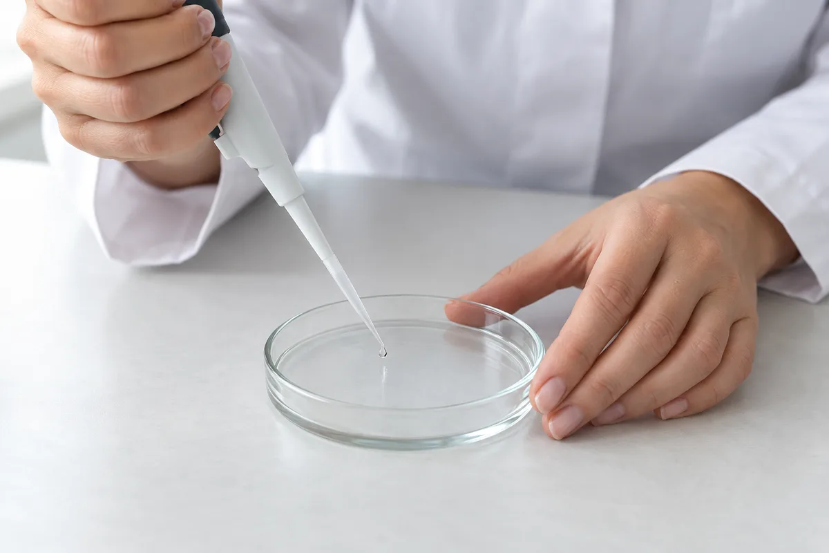 Mains d&rsquo;une femme en blouse blanche déposant une goutte de pipette dans une boîte de Petri sur une paillasse claire
